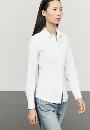 Femme aux longs cheveux noirs portant une chemise blanche boutonnée et un jean bleu clair, marchant devant un fond gris clair uni.