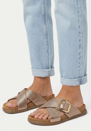 Füße in hellblauen hochgekrempelten Jeans und metallisch roségoldenen Sandalen mit gekreuzten Riemen, Korksohle und Schnallendetail.