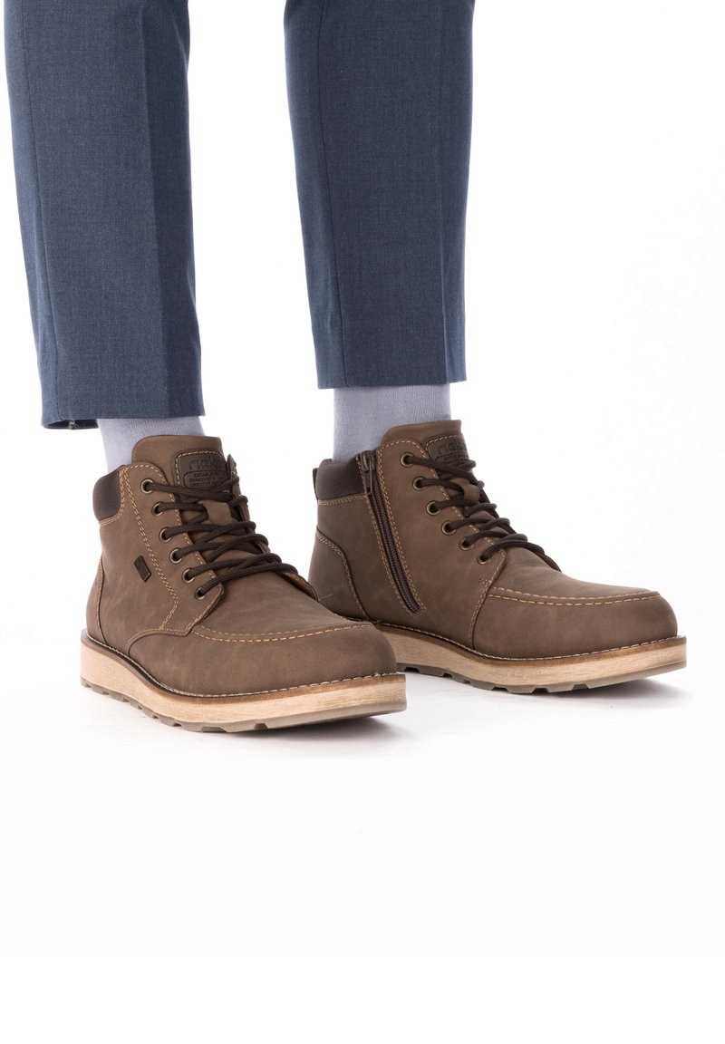 Brown leather ankle boots with a textured finish, stitched accents, and a side zipper, paired with gray socks and tailored blue trousers.