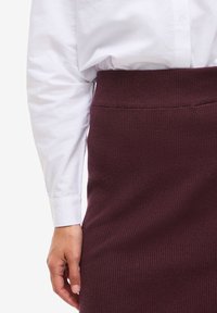 Burgundská žebrovaná sukně s měkkou texturou a širokým pasem, spárovaná s bílou košilí s knoflíky a dlouhými rukávy.