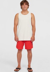 Homme debout sur fond blanc portant un débardeur gris clair, un short de bain rouge avec un logo noir, et des tongs noires.