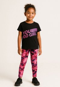 Young girl standing, wearing a black t-shirt with "YOU GO GIRL" in pink and pink patterned leggings with black sneakers.
