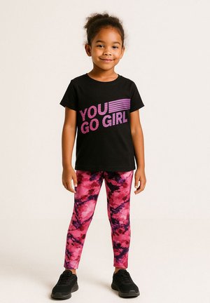 Jong meisje staat, draagt een zwart T-shirt met "YOU GO GIRL" in het roze en roze legging met patronen en zwarte sneakers.