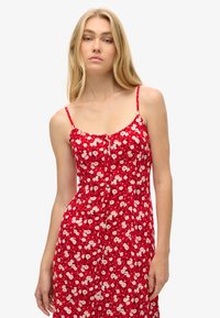 Rotes Blumenkleid mit dünnen Trägern, Knopfdetails und einer taillierten Passform. Mit kleinen weißen und rosa Blumen auf einem einfarbigen roten Hintergrund.