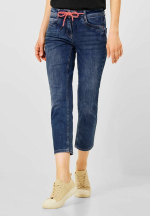 Person iført blå korte jeans med en pink snor og beige sneakers, stående på en hvid baggrund.