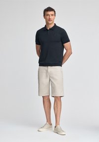 Camisa polo negra con tres botones, pantalones cortos beige y zapatillas de color claro. Modelo de pie con las manos detrás de la espalda, fondo neutro.