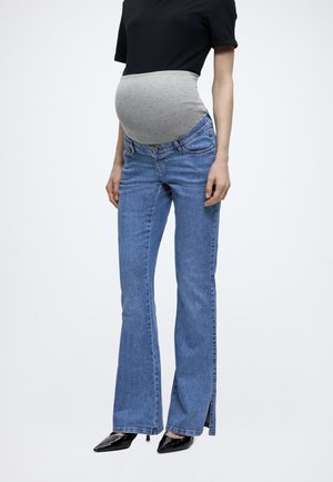 Tehotná osoba oblečená v modrých tehotenských džínsoch so šedým pružným pásom, čiernom topu a čiernych špicatých podpätkoch na bielom pozadí.