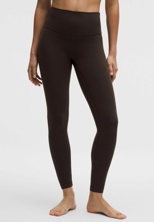 Mujer con leggings negros de cintura alta y longitud total, de pie descalza sobre un fondo claro.