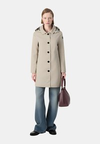 Cappotto impermeabile beige con cappuccio, chiusura frontale con bottoni e tasche laterali. Indossato con jeans in denim a gamba larga e una borsa bordeaux.
