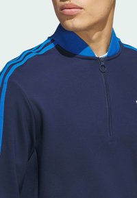 Navy zip-up jacket with blue ribbed collar and three blue stripes along shoulders. Smooth fabric with a slim fit design.