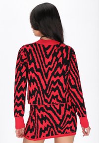 Pulover tricotat roșu și negru cu un model în zigzag, având guler rotund și manșete îndoite, prezentat din spate. Textură netedă.