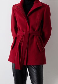 Abrigo rojo de mezcla de lana con cuello alto, cierre de cremallera y cintura ceñida. Cuenta con dos bolsillos laterales y una forma entallada. Combinado con pantalones negros acolchados.