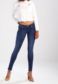 Vit kortärmad skjorta med blommig brodyr, kombinerad med mörkblå tajta jeans och ljusbeige högklackade skor.