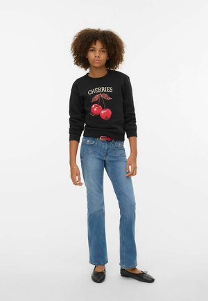 Junge Person mit lockigem Haar trägt einen schwarzen "CHERRIES"-Sweatshirt, blaue Jeans mit einem roten Gürtel, schwarze Ballerinas und ein Armband.