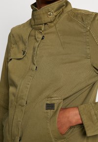 Olivově zelená bunda s vysokým límcem, zapínáním na přední druky a bočními kapsami. Má texturovanou tkaninu a nášivku s logem na kapse.