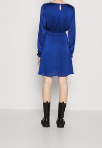 Robe en satin bleu à manches longues, avec une taille élastique et une ouverture en forme de trou de serrure dans le dos, associée à des bottines noires.