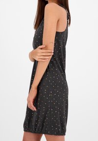 Kobieta w bezrękawowej, ciemnoszarej sukience z kolorowym, drobnym wzorem liści, trzymająca lewą rękę, widoczny tatuaż na ramieniu, na białym tle.