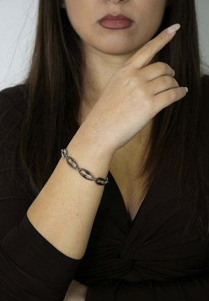 Bransoletka z łańcuszkiem ogniwowym w ciemnych metalicznych odcieniach, z owalnymi oczkami, noszona na jasnej skórze. Gładka tekstura, minimalistyczny design.