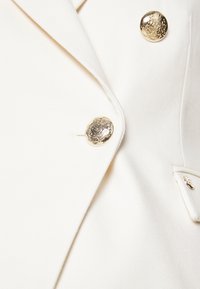 Primo piano di un blazer bianco in tessuto con bottoni metallici dorati incisi e una patta sul taschino.