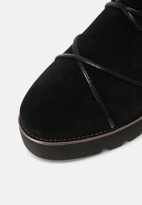 Chaussure en cuir suédé noir avec un bout arrondi, ornée de brides croisées noires brillantes et d'une semelle en caoutchouc épaisse avec un discret détail de couture contrastante.