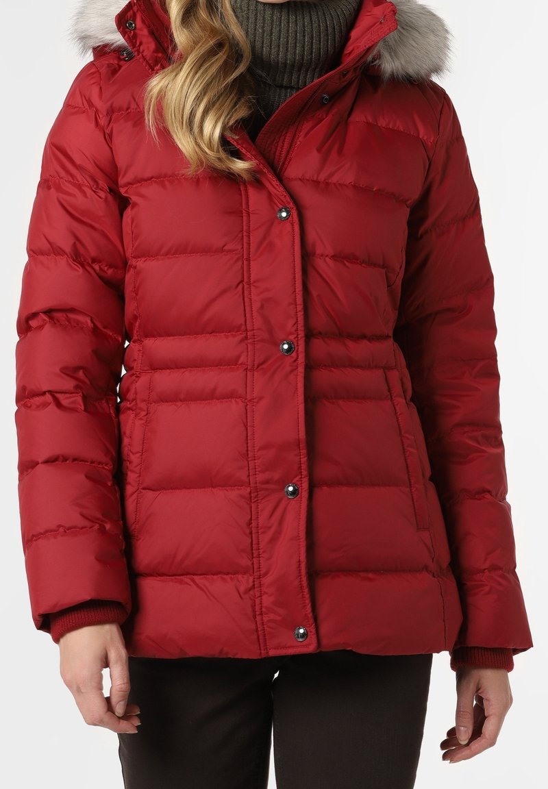 Femme portant une veste d'hiver matelassée rouge avec capuche bordée de fourrure et un pull à col roulé foncé, debout les mains détendues le long du corps.