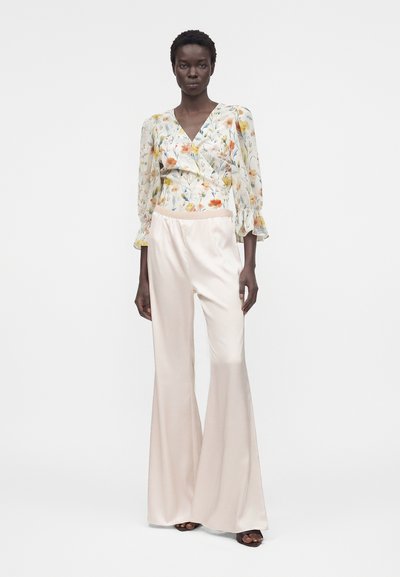 Γυναίκα με φλοράλ μακρυμάνικη μπλούζα με V-λαιμόκοψη και κρεμ σατέν παντελόνια με φαρδύ πόδι, στέκεται με ουδέτερη έκφραση.