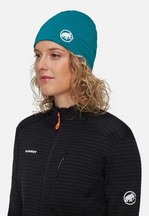 Berretto elastico turchese con una texture liscia e un logo di un elefante bianco. Indossato con una giacca nera con zip caratterizzata da strisce orizzontali.