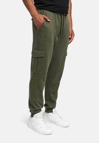 Man in olijfgroene cargo jogger pants met een broekkoord in de taille en witte Nike sneakers, met handen in de zakken, tegen een witte achtergrond.