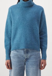 Pull à col roulé bleu en tissu doux et duveteux. Comporte un col et des poignets côtelés, avec une coupe décontractée et un ourlet légèrement raccourci.