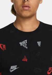 T-shirt nera in cotone con vari logo Nike e il testo "Just Do It" in rosso e bianco, con una vestibilità regular e maniche corte.