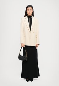 Woman wearing a cream blazer over a black dress with beige piping, holding a black handbag, standing against a plain white background.