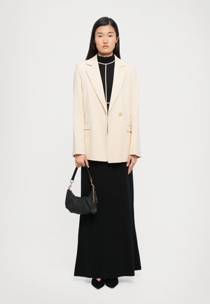 Woman wearing a cream blazer over a black dress with beige piping, holding a black handbag, standing against a plain white background.