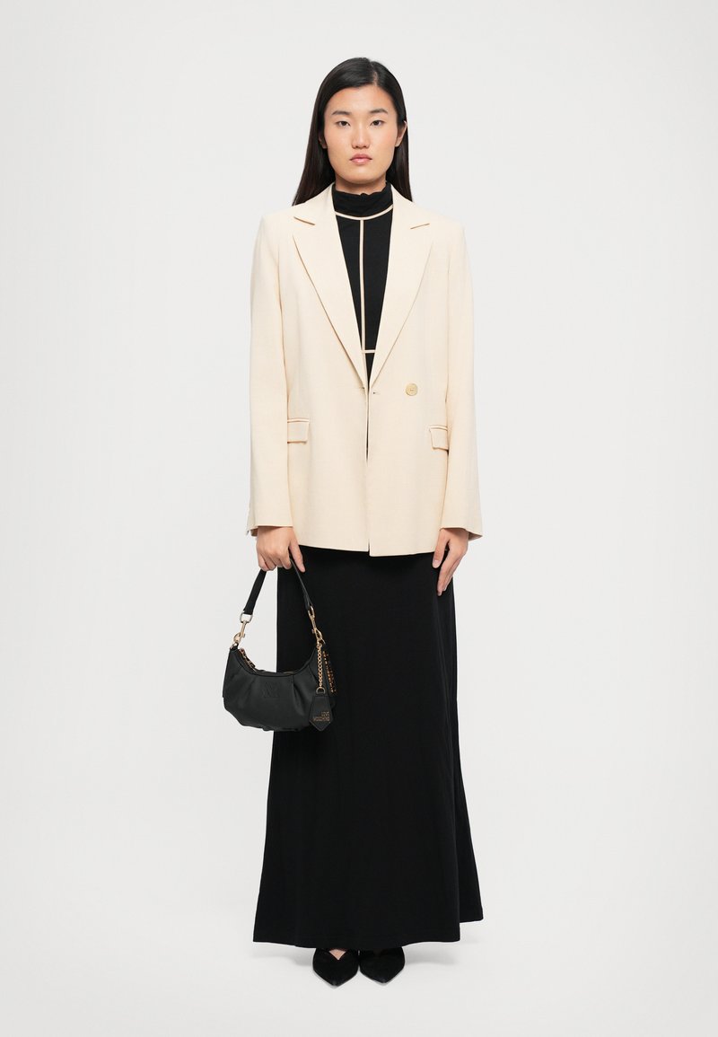 Woman wearing a cream blazer over a black dress with beige piping, holding a black handbag, standing against a plain white background.