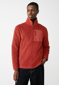 Roter Fleece-Pullover mit hohem Kragen, Halb-Reißverschluss auf der Vorderseite und einer Brusttasche in passender Farbe. Weiche Textur, lässige Passform.