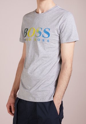 Mężczyzna noszący jasnoszarą koszulkę z krótkim rękawem z wielokolorowym napisem "BOSS HUGO BOSS" i ciemne spodnie, stojący z jedną ręką w kieszeni.