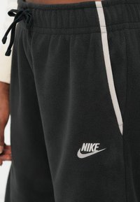 Črne flis jogger hlače z teksturirano površino, ki imajo belo stransko črto in srebrni Nike logotip na spodnjem levem nogavicu. Prilagodljiv pas.