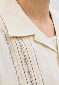 Camisa de crema clara con cuello y rayas verticales en rojo, naranja, azul y verde, y bordados intrincados a lo largo del frente.