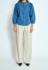 Džínová košile na knoflíky s nafouknutými rukávy, doplněná béžovými širokými kalhotami a černými lodičkami s špičatou špičkou. Kulatý límec a viditelné knoflíky.