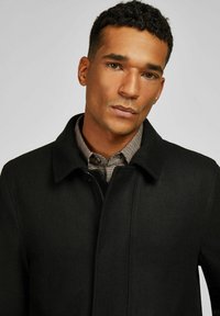 Man met kort krullend haar in een zwarte jas over een bruine geruite overhemd, recht vooruit kijkend met een neutrale uitdrukking.