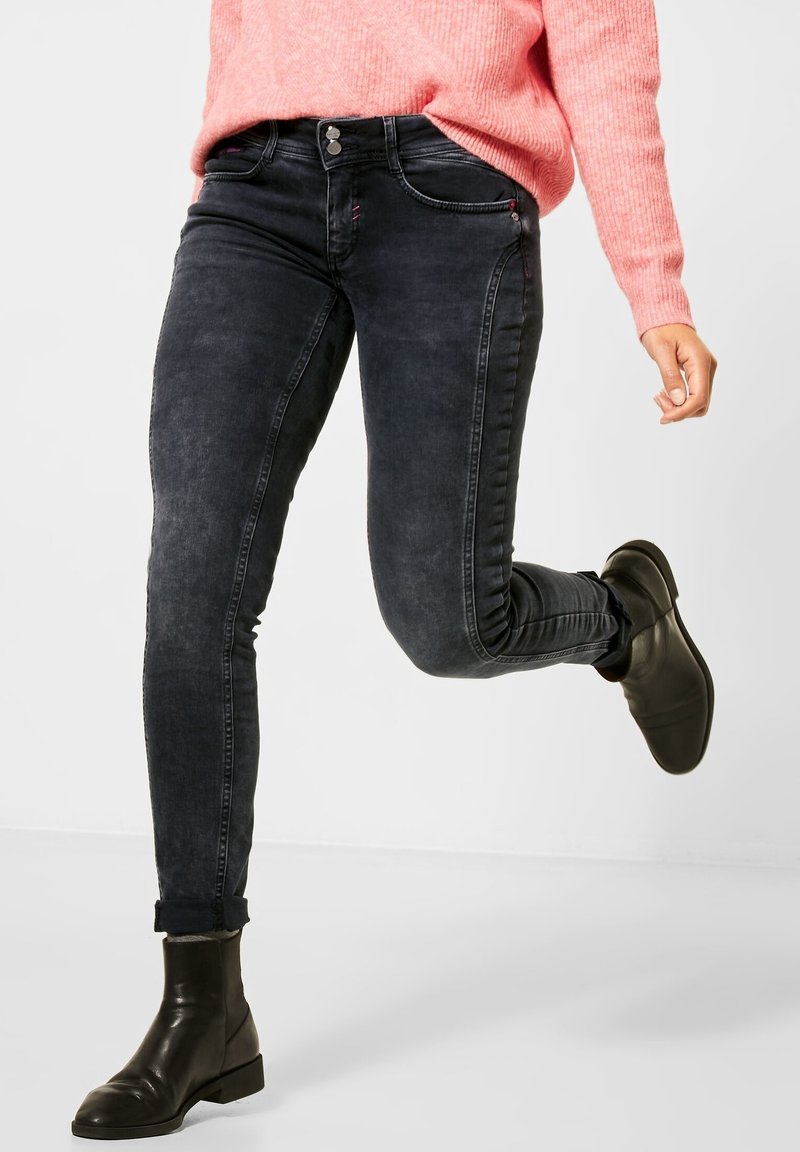 Personne portant un jean noir slim, des bottines noires et un pull rose, levant une jambe comme à mi-pas devant un fond uni.
