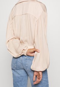 Henkilö, joka pukeutuu vaaleanbeigeen, löysästi istuvaan yläosaan, jossa on puhvihihat, seisoo näyttäen vaatteen selkäpuolta yhdistettynä sinisiin farkkuihin.