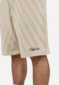 Shorts en tricot beige avec des rayures verticales, présentant des motifs ajourés. Comprend un brodage de logo noir à l'ourlet. Fabriqué à partir de matériau doux.