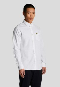 Camisa blanca de manga larga hecha de una tela suave con cuello abotonado, que presenta un pequeño logo de mariposa amarilla en el bolsillo izquierdo del pecho.