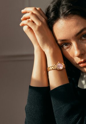 Tummatukkainen nainen lepää kasvojaan kiedotuissa käsissään ja käyttää kultakelloa, jossa on vaaleanpunainen kellotaulu, ranteessaan.