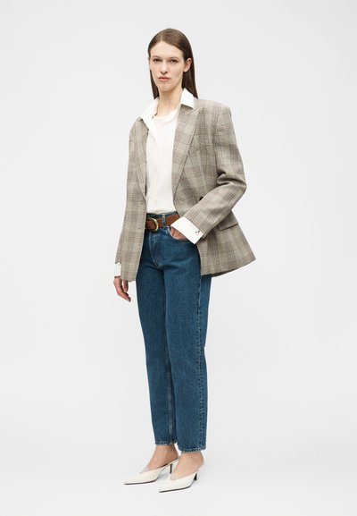 Frau trägt einen karierten beige Blazer, weißes Hemd, blaue Jeans mit braunem Gürtel und weiße, spitze Kitten-Heels, steht vor einem schlichten Hintergrund.