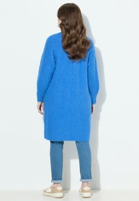 Langer blauer Pullover aus flauschigem Material, mit geradem Schnitt und geripptem Saum, getragen über hellblauen Jeans und beigefarbenen Sneakern.