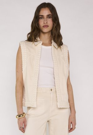 Femme aux cheveux bruns ondulés portant un gilet tissé sans manches de couleur blanc cassé, un débardeur blanc, un pantalon assorti, un bracelet en or et des bagues, sur un fond uni.