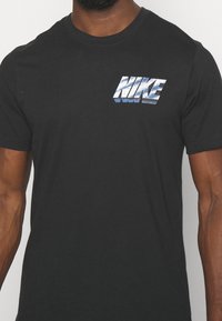 Černé bavlněné tričko s reflexním stříbrným logem "NIKE" na horní levé straně. Krátké rukávy, kulatý výstřih, jemná textura.