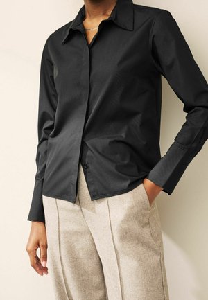 Personne portant une chemise noire à manches longues avec col, rentrée dans un pantalon beige à chevrons taille haute, main dans la poche, sur un fond uni.