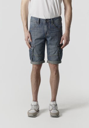 Hombre con shorts cargo de mezclilla azul remangados, zapatillas blancas y camiseta blanca, de pie contra un fondo claro y liso.